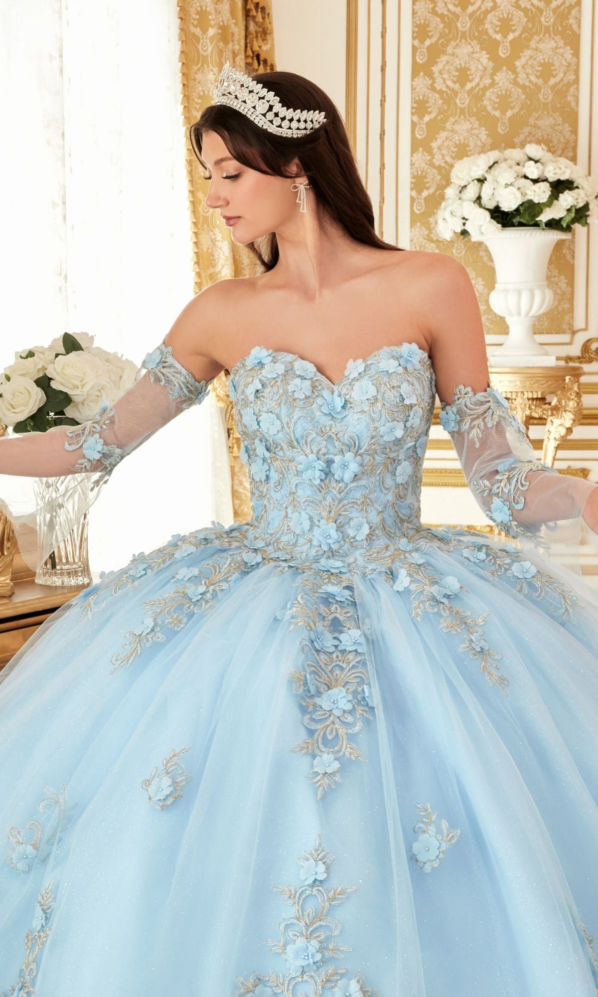 A woman wears the Quinceanera Dress 15714 by Ladivine, a pastel ball gown with lace embroidery and sheer sleeves, paired with a jeweled tiara. She stands indoors, surrounded by white roses and ornate gold décor.